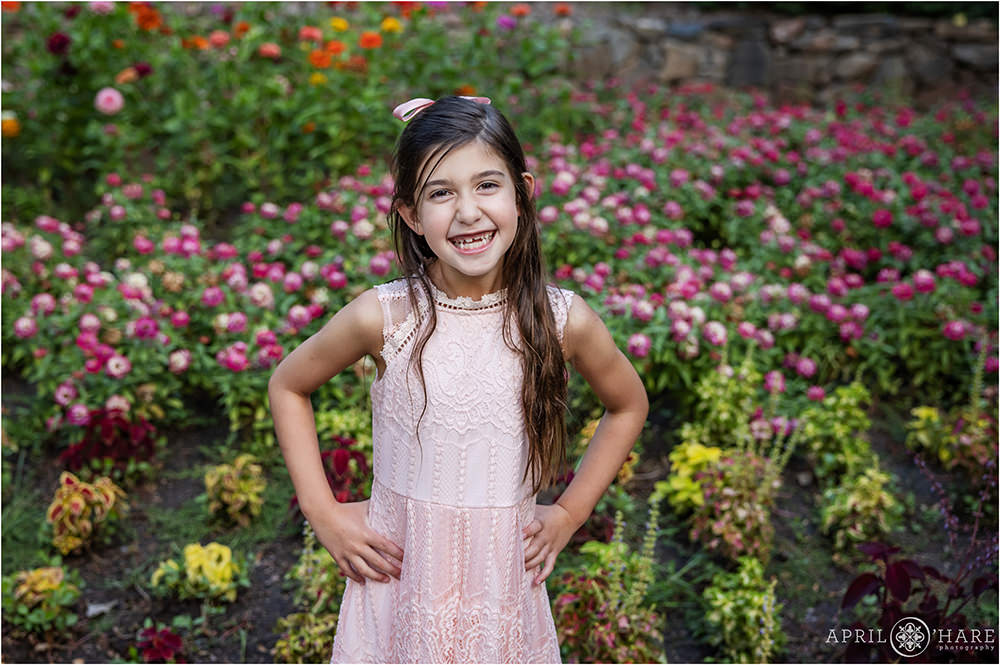 Cute girl with long brown hair wearing a pink ribbon and a light pink lace dress poses with her hands on her hips and smiles big in front of the zinnia flowers at Gallup Gardens in Littleton Colorado