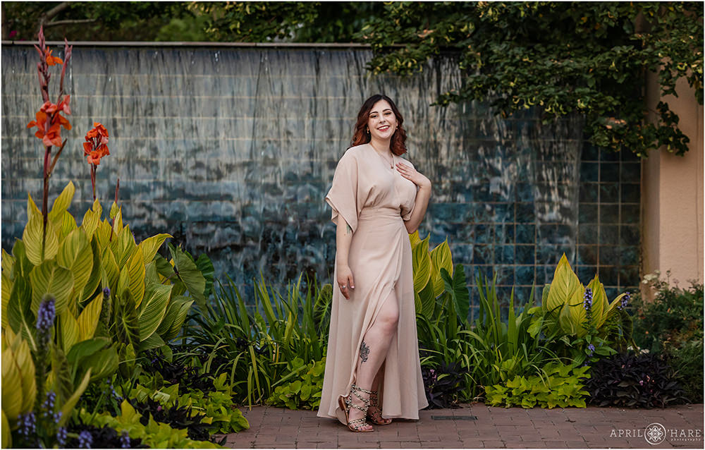 A full length high school senior portrait for a young lady with ombre red hair wearing a light pink floor length gown in front of the blue tile waterfall fountain at Denver Botanic Gardens