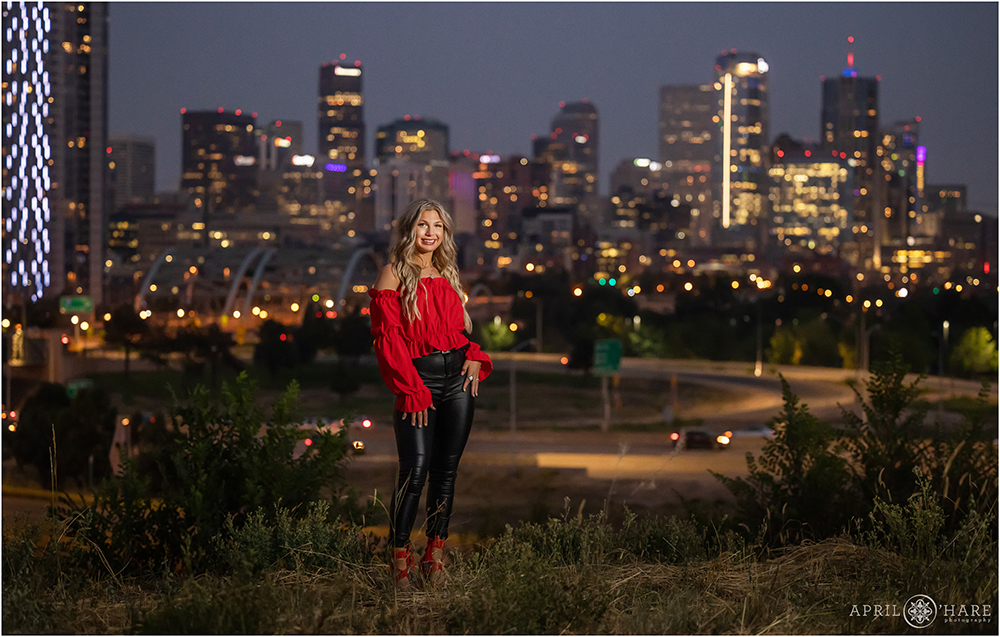 Night time portrait for a high school senior girl with long blonde hair wearing a red flowy top with leather pants standing in front of the Denver skyline lit up at dusk