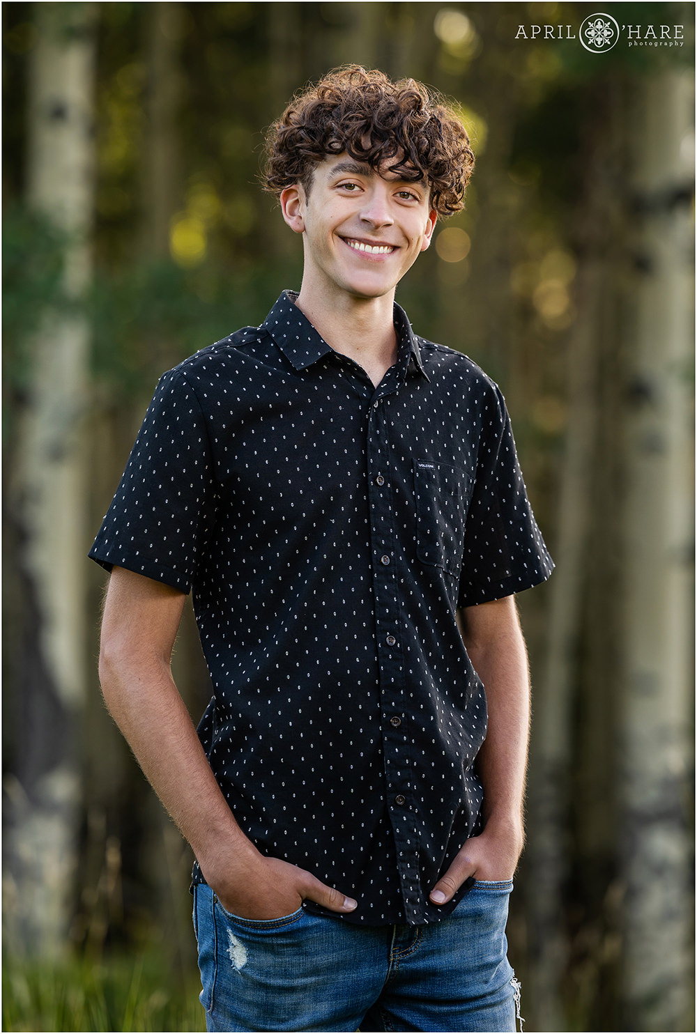 Senior boy with curly brown hair wearing a dark shirt with a dotted print on it in the woods of Colorado