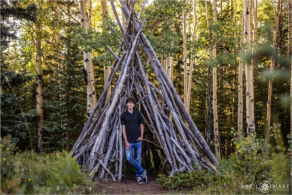 High school senior boy stands with a wood tipi in the woods of Colorado