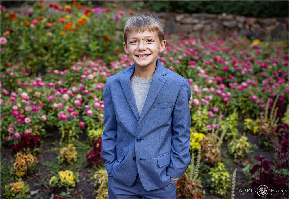Cute boy with blonde hair wearing a blue suit smiles big in front of the flower garden at Gallup Gardens in Littleton Colorado