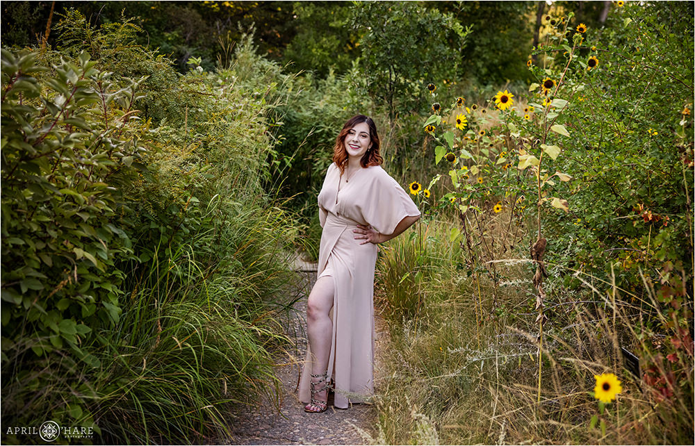 A senior girl stands on a path wearing her floor length light pink gown in the long grasses and sunflower area of Denver Botanic Gardens
