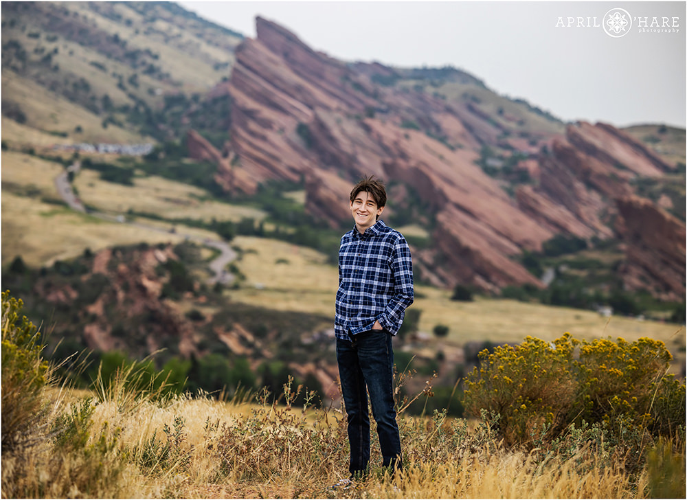 Dramatic scenery of Red Rocks Amphitheater provides a really cool backdrop for a full length senior photo for a dark haired boy wearing dark blue jeans and a blue plaid long sleeved button down shirt