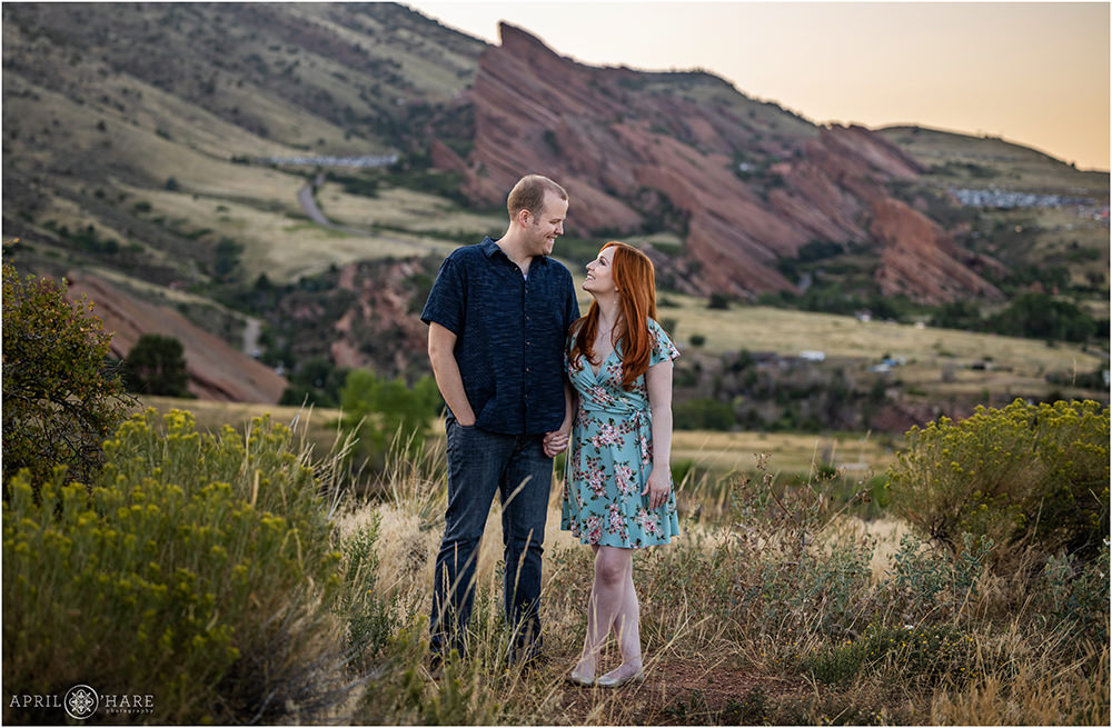 A couple holds hands and gazes into each other's eyes with plants growing around them at East Mount Falcon Trailhead with views of Red Rocks Amphitheater behind them