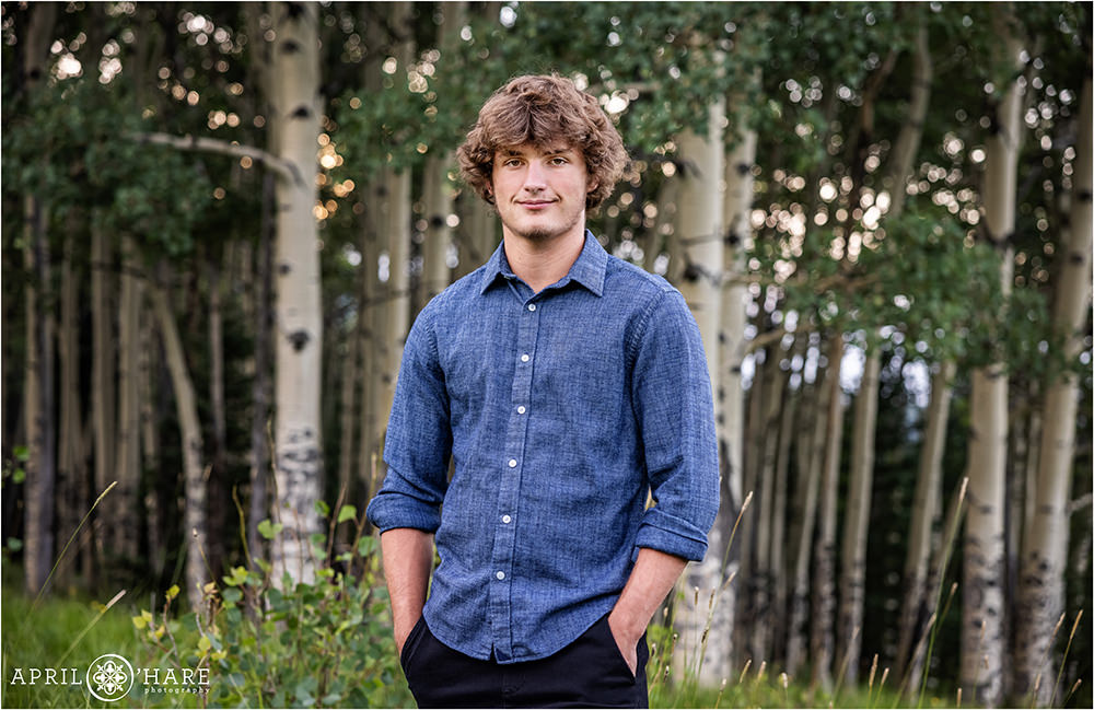 High school senior portraits for a boy wearing black pants and a blue long sleeved button up shirt with sleeves rolled up standing in a mountain meadow with aspen trees in the backdrop