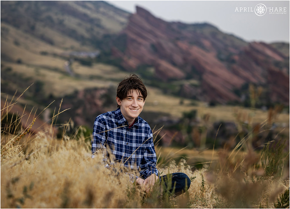 A high school senior boy sits criss cross style on the ground at the East Mount Falcon trailhead area with Red Rocks off in the distance behind him