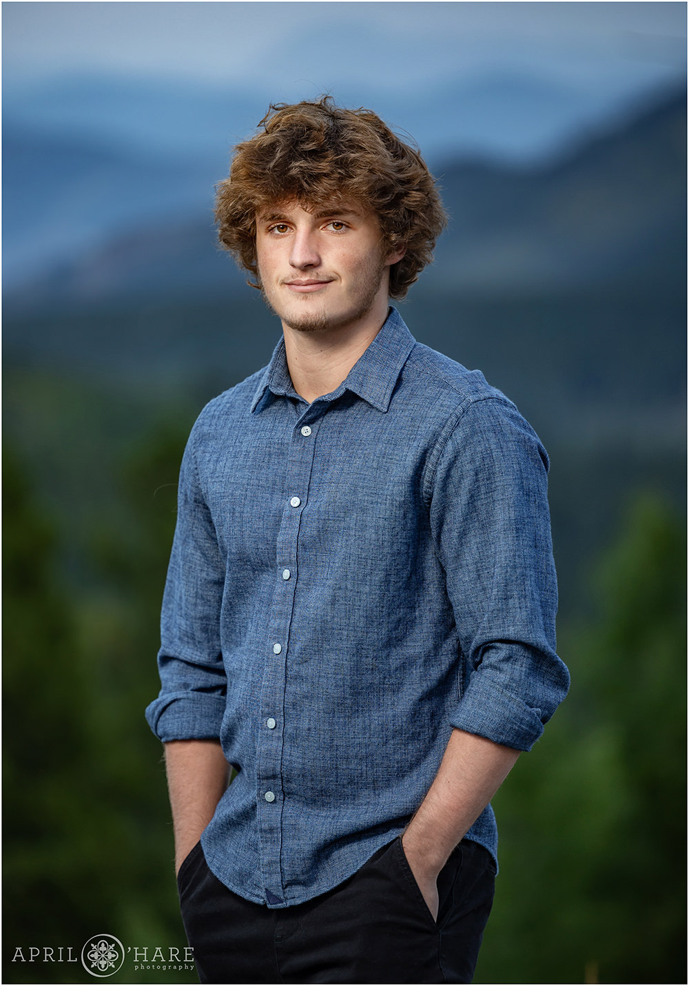 High school senior boy wearing a slong sleeved blue chambray button down shirt with the sleeve rolled up yearbook photo with a nice hazy mountain backdrop