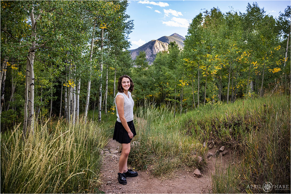 A high school senior girl with shoulder length brunette hair wearing a black skirt and a white sleeveless sweater top poses on the Meadow Creek trail with aspen trees and a mountain backdrop behind her