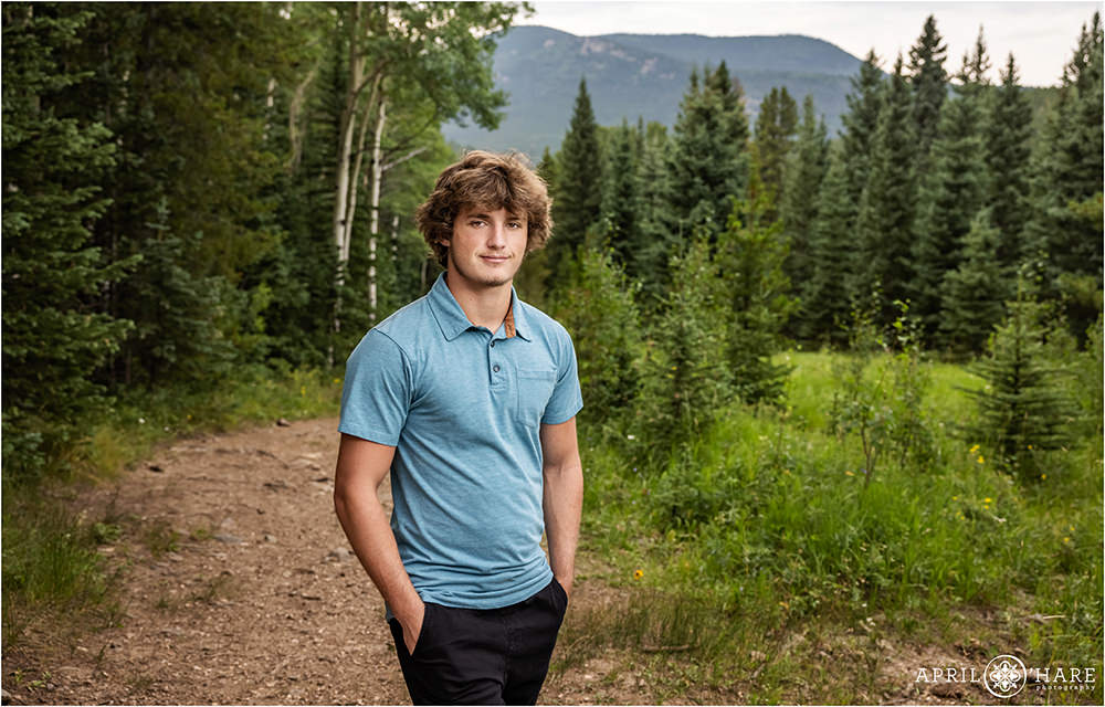 High school senior boy with light brown hair wearing a light blue shirt stands on a trail in the mountain of Evergreen Colorado