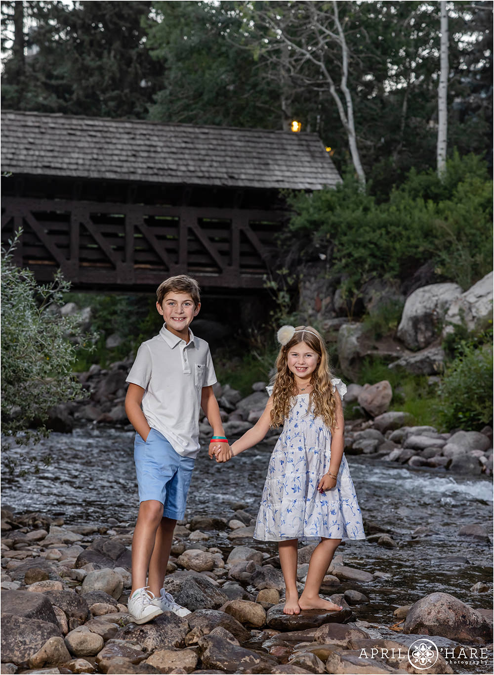 Brother and sister wearing blue and white hold hands in front of the Covered Bridge along Gore Creek in Vail during summer family portraits