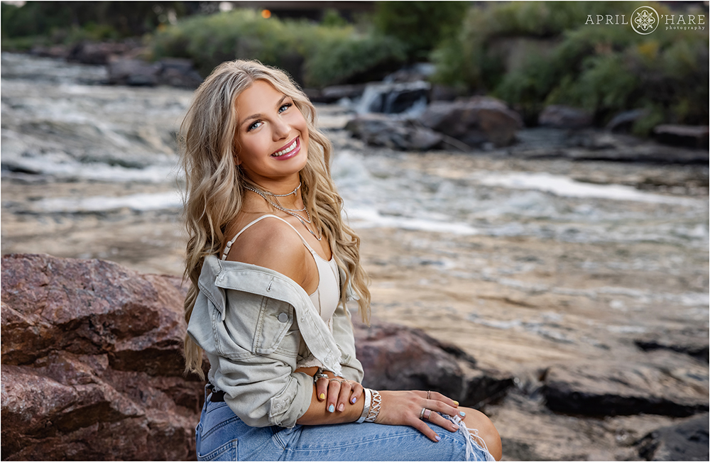 High school senior girl with long blonde hair sits next to the South Platte River across from REI and the confluence with Cherry Creek for her high school senior yearbook photos in downtown Denver