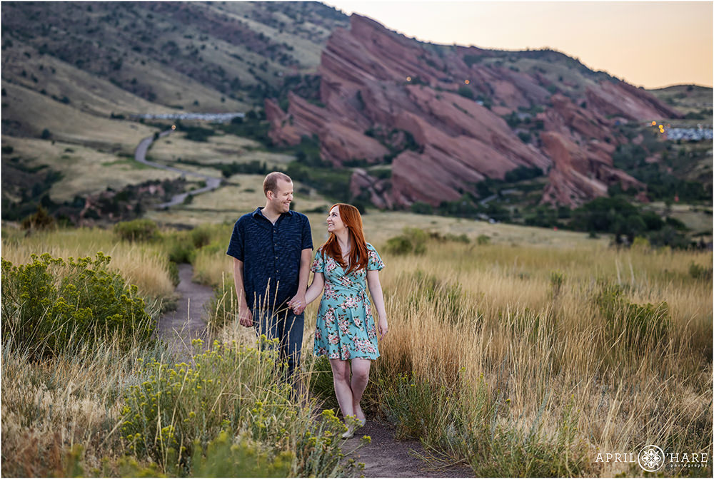 Couple walk hand in hand down the path at East Mount Falcon at sunset with Red Rocks Amphitheater off in the distance