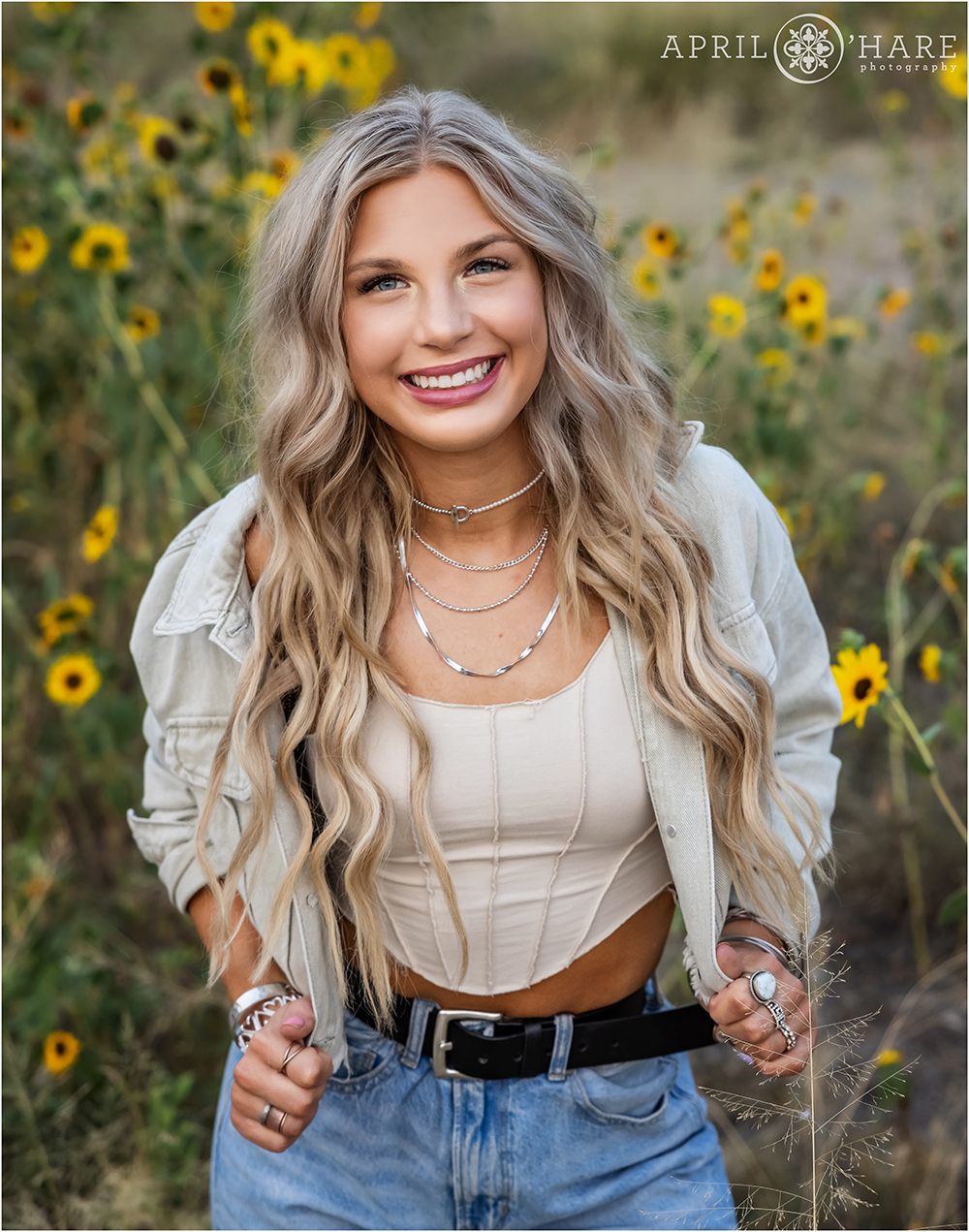 A high school girl with long blonde wavy hair wearing blue jeans and a tan jean jacket poses in front of some pretty sunflowers in Denver Colorado