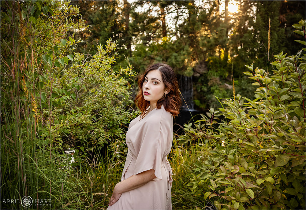A high school senior girl with reddish brown hair wearing a light pink dress poses in front of a small waterfall at Denver Botanic Gardens