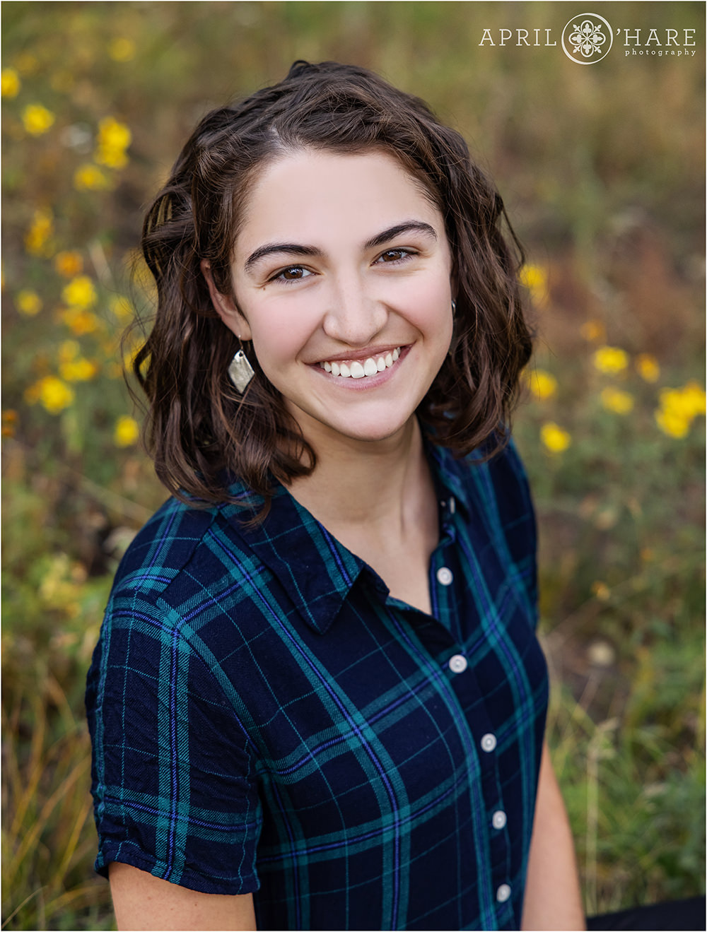 A brunette high school senior wearing silver earrings and a blue and black plaid short sleeve top poses in a small strand of yellow wildflowers at her Frisco Colorado senior session