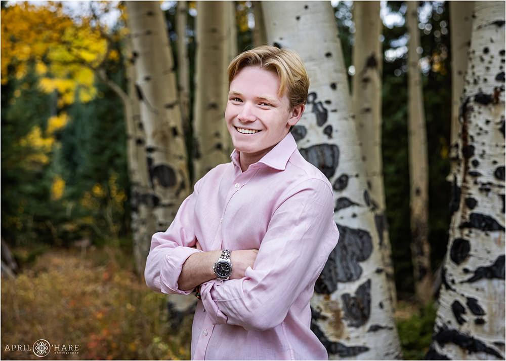 Young senior boy with blonde hair wearing a pink button down shirt leans against an aspen tree with fall color in the backdrop