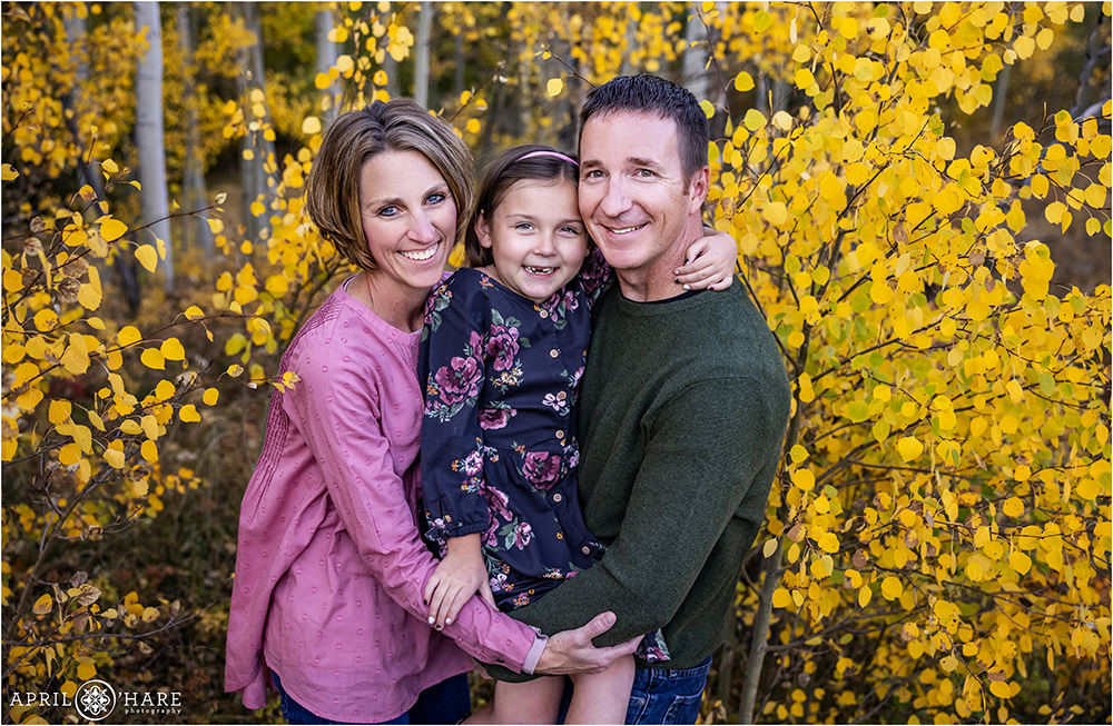 A beautiful photo of a family of 3 with a young daughter held between them surrounded by golden aspen leaves during fall in Colorado