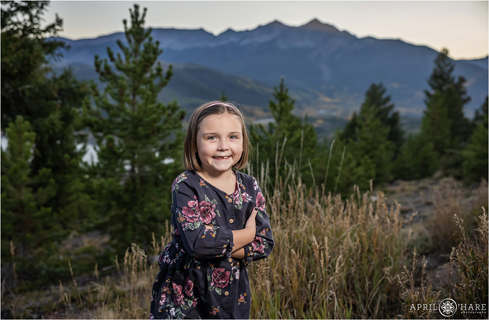 A cute little girl with her arms crossed waring a dark floral dress and pink headband smiles at Sapphire Point at her mountain family session in Colorado