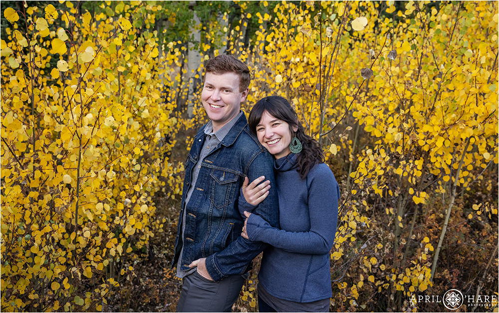 A couple wearing blue stand in a grove of aspens with bright yellow color during fall in Colorado