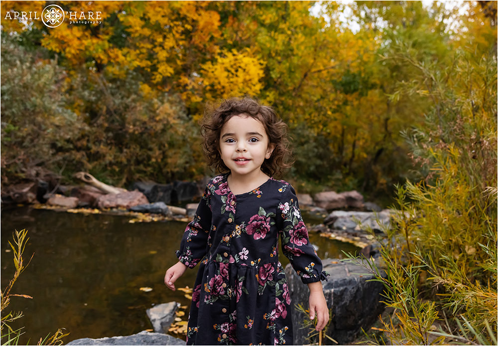 Adorable little girl with curly brown hair stands in front of a creek at James A. Bible Park in Denver CO