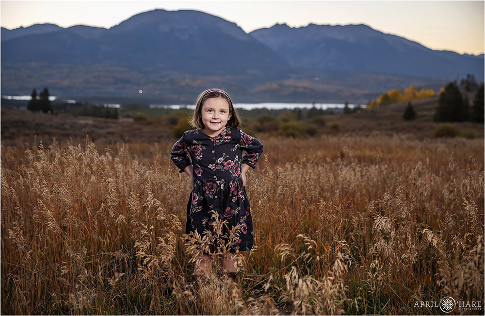 A little girl with short shoulder length hair stands with her hands on her hips with a pretty fall color mountain backdrop in the distance