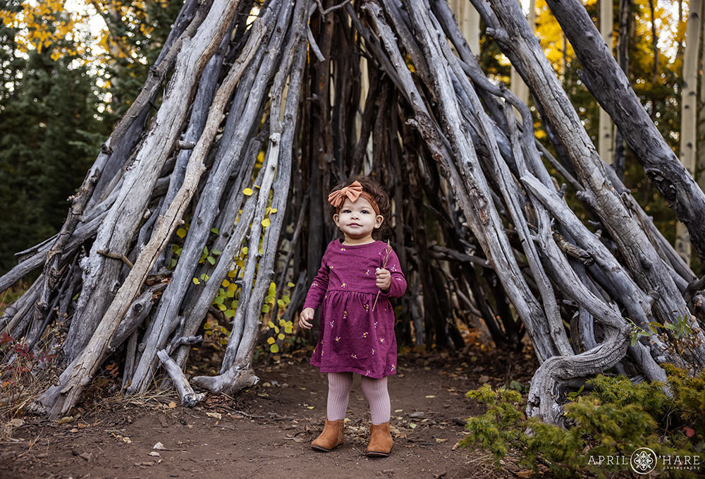 Cute toddler wearing a plum colored dress with little yellow flowers on it stands in front of a wood tipi with a bow in her hair and wearing brown booties