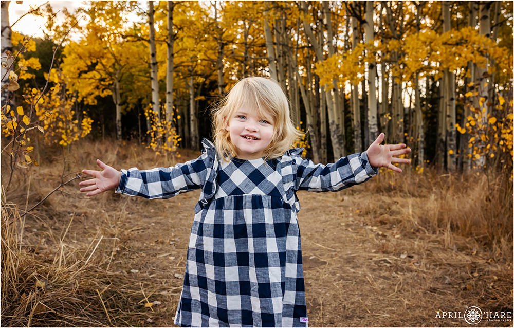 Sweet photo of a toddler girl with blonde hair wearing a blue and white checkered dress in a golden aspen tree forest in Colorado
