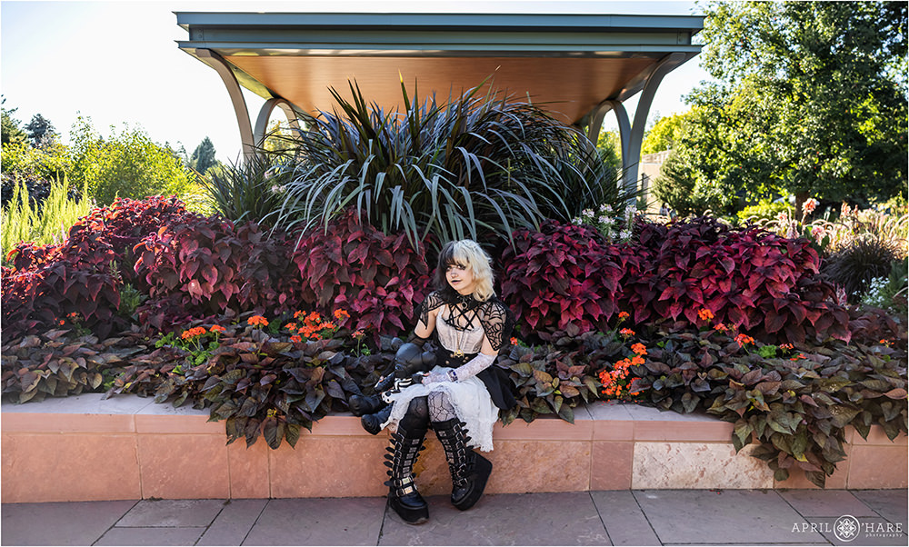 Senior girl wearing a black sheer cape and B&W clothing sits in front of a dark plant garden at Denver Botanic Gardens