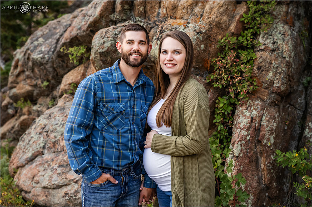 Married couple hold hands as they pose for a pregnancy portrait together in front of a rocky backdrop at Lily Lake in Estes Park Colorado