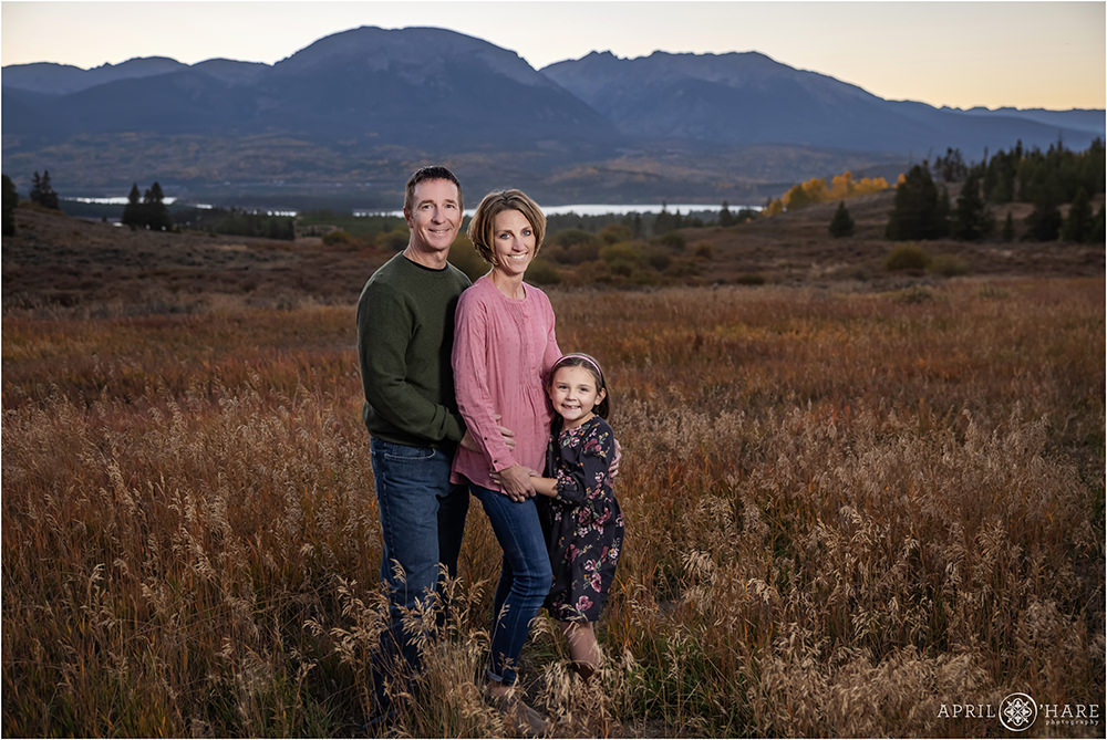 A family of 3 with a young daughter wearing a plum colored floral dress poses together with Buffalo Mountain in the backdrop in Summit County Colorado
