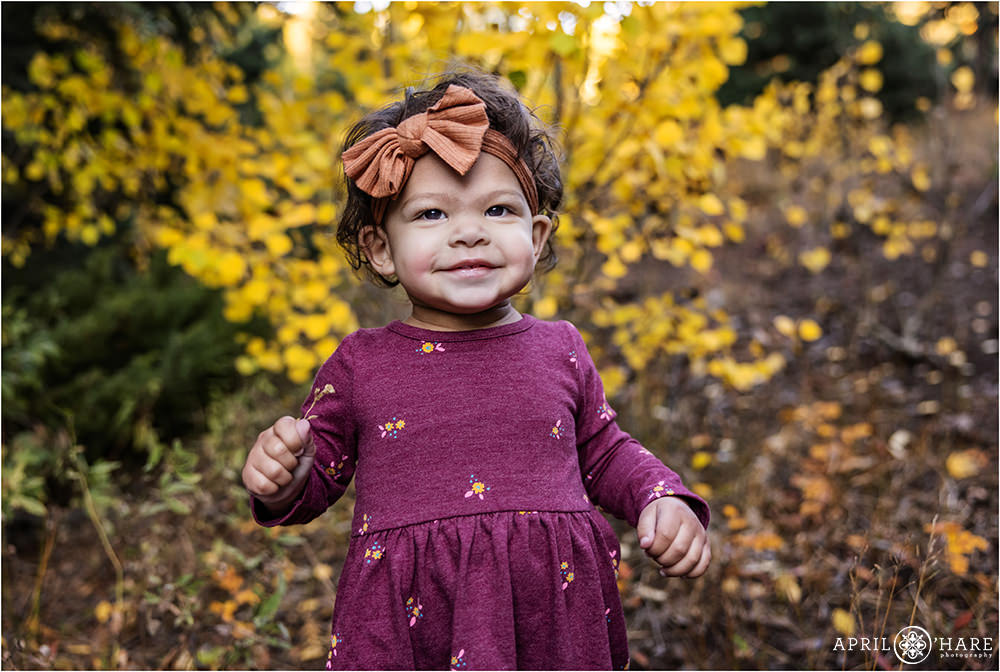 A sweet little toddler girl with a bow in her hair smiles for the camera in front of a fall color backdrop in Evergreen Colorado