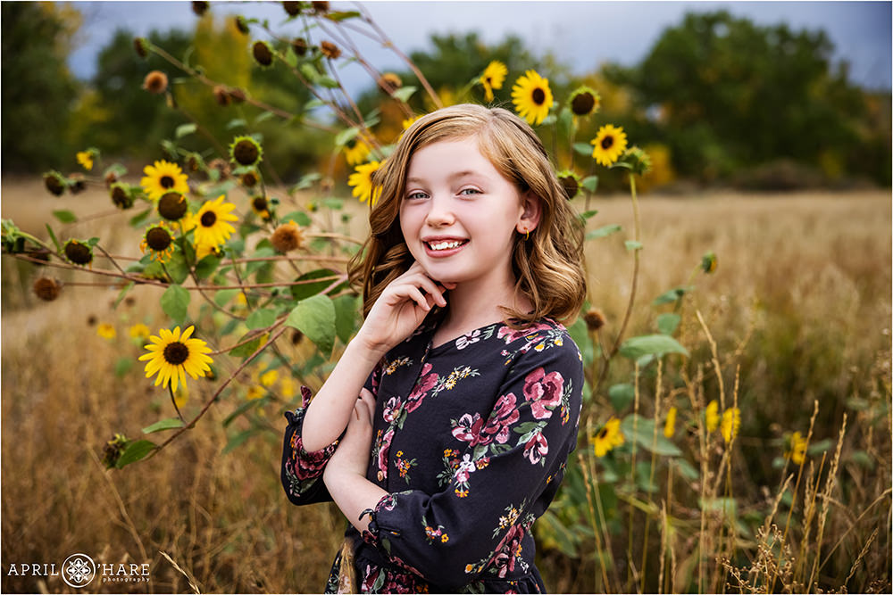 Tween girl with reddish blonde hair wearing a dark floral dress in the wild sunflowers at James A. Bible Park in Denver Colorado