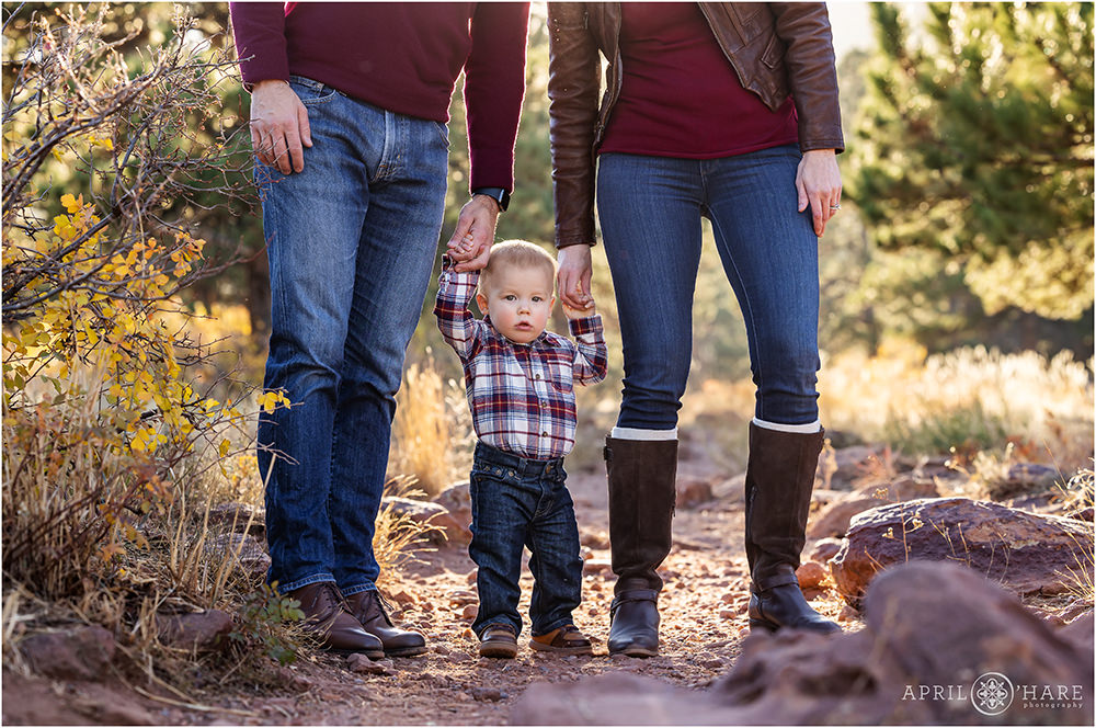 Photo of a young baby boy standing on a path holding his parents hands during their family portrait session in Boulder