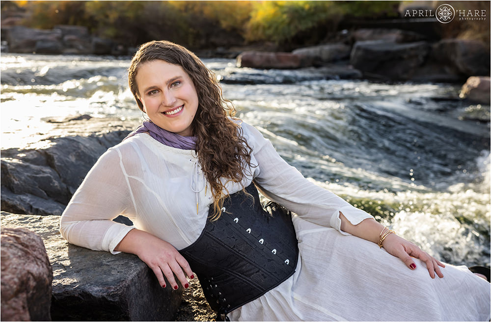 Woman with long curly hair wearing a flowy white dress with a black corset poses in front of the South Platte River in Denver Colorado