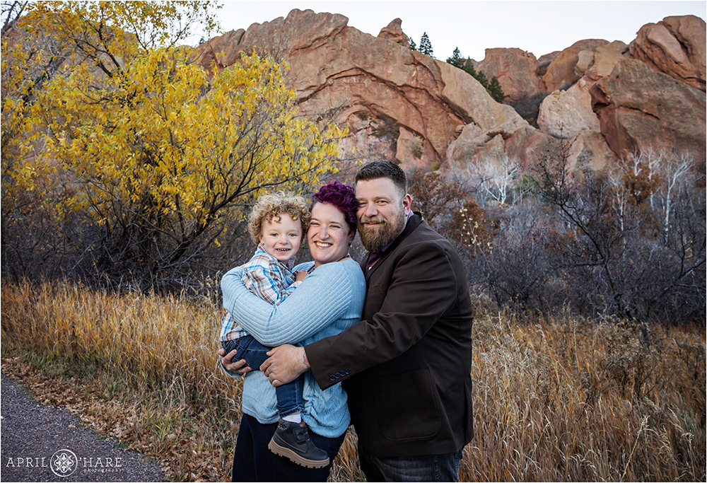 A family of three cuddle together with fall color backdrop at Roxborough State Park in Colorado