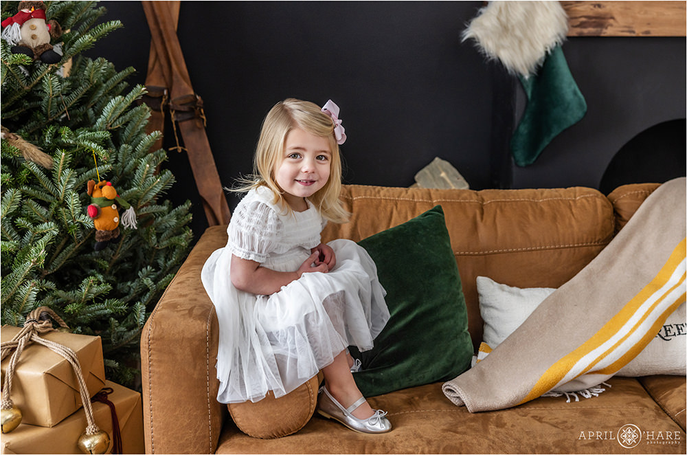 Little Girl wearing a white dress with a pink bow in her blonde hair poses on a mid century modern style couch in a styled Christmas setting at Denver Photo Collective