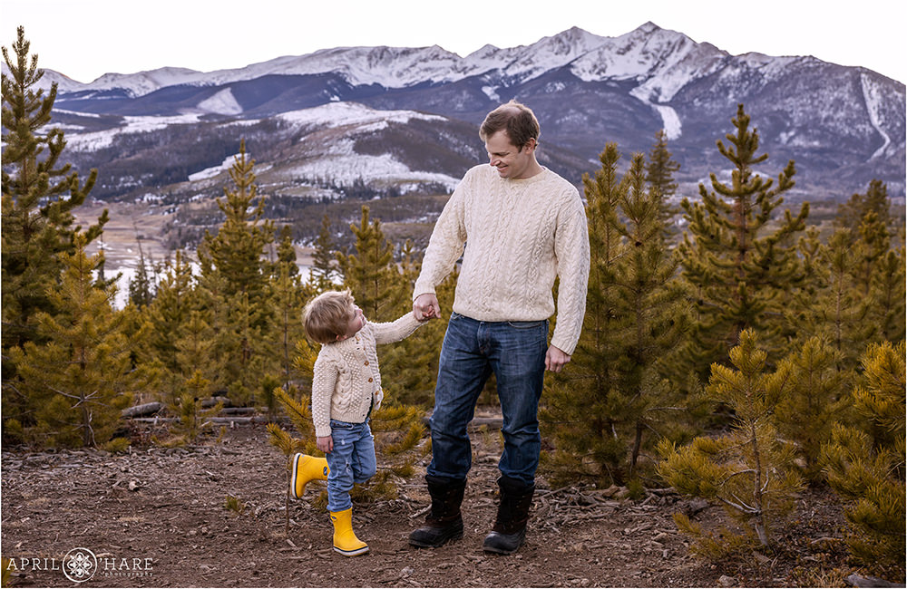 A young son and his dad wearing yellow boots and cream sweaters hold hands in front of the pretty Sapphire Point mountain view in Summit County Colorado