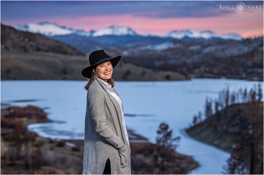 Woman wearing a western style hat and gray cardigan stands in front of a beautiful pink and purple sunset at C Lazy U Ranch in Colorado