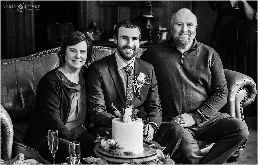 B&W portrait of groom with his parents sitting on sofa with his cute white wedding cake at Romantic Riversong Inn in Estes Park Colorado