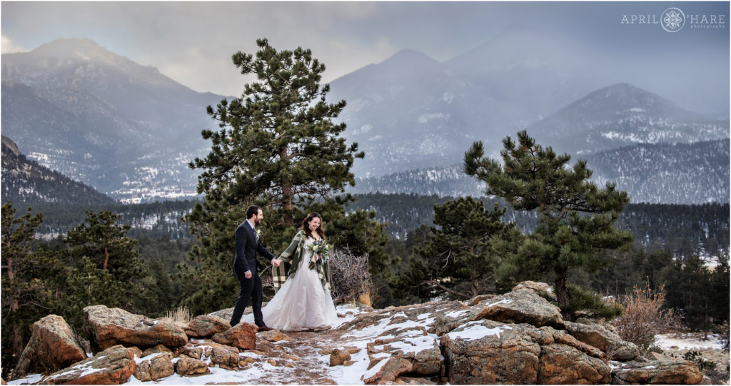 Bride and groom walk along a rocky path with a beautiful snowy winter mountain landscape in the backdrop at Rocky Mountain National Park