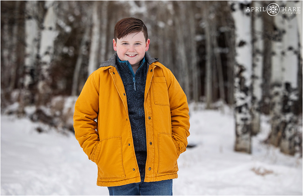 Cute boy wearing a bright yellow jacket standing in a pretty winter aspen tree forest in Beaver Creek Colorado