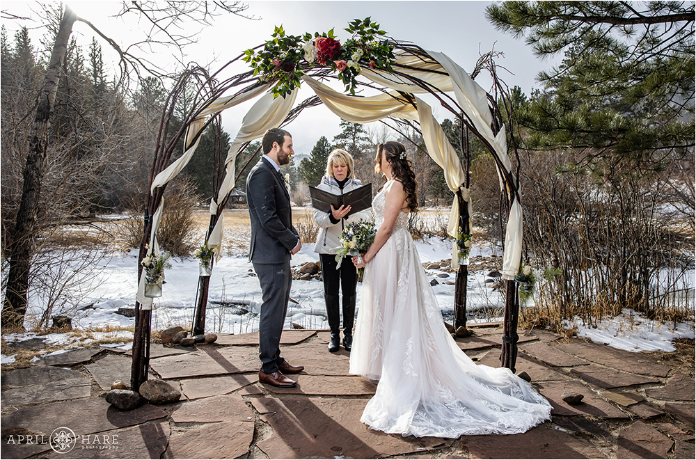 Couple get married outside in front of a tree branch arch decorated with white fabric and florals on their winter wedding day at Romantic Riversong Inn in Colorado