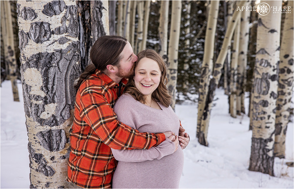 Father to be kisses his pregnant wife at their maternity photoshoot in an aspen forest during winter in Colorado