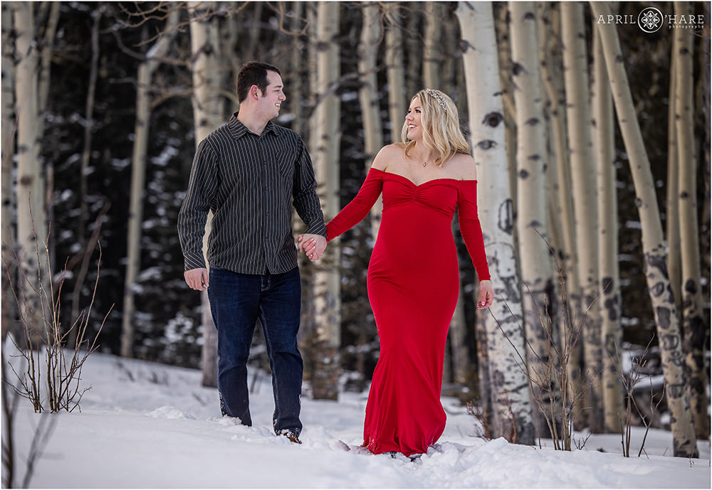 Snowy maternity portraits in an aspen tree forest with a mama to be wearing a long flowy red dress in Colorado
