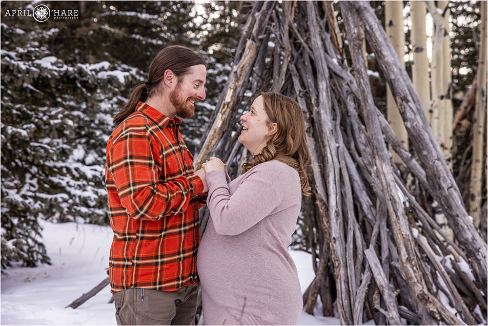 Couple holds hands and look at each other in a winter forest setting during their maternity session