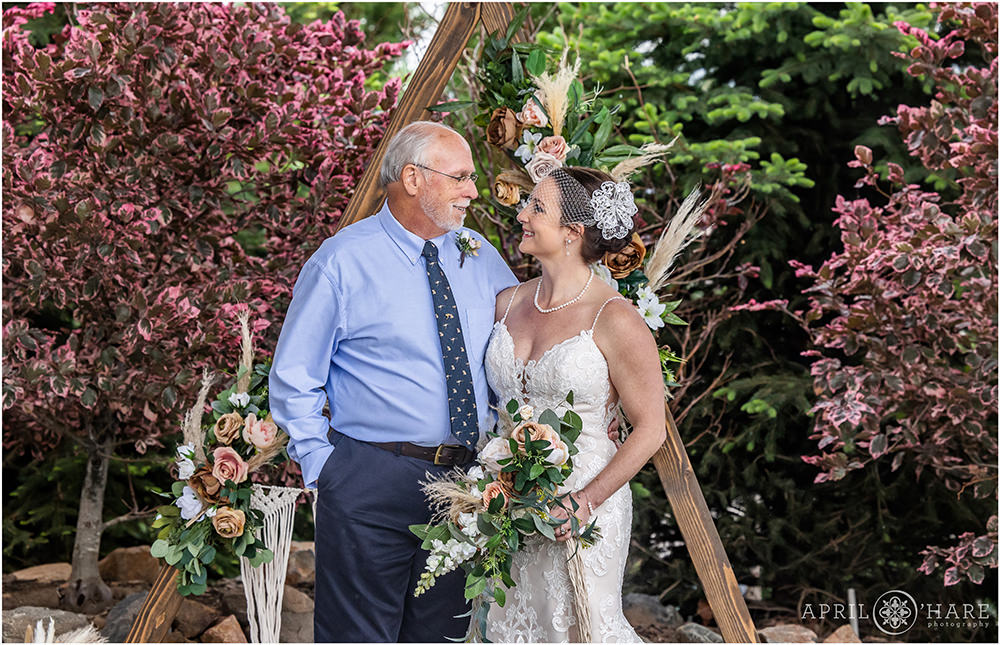 A woman and her dad smile at each other in front of a pretty floral boho triangle wood arch at her 20th wedding anniversary vow renewal party in Colorado