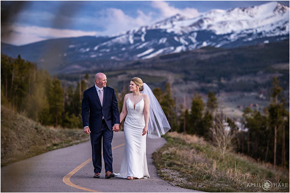 Bride and groom walk hand in hand down the bike path next to Sapphire Point with the Breckenridge Mountains in the distance in Colorado at their sunset summer wedding in Colorado