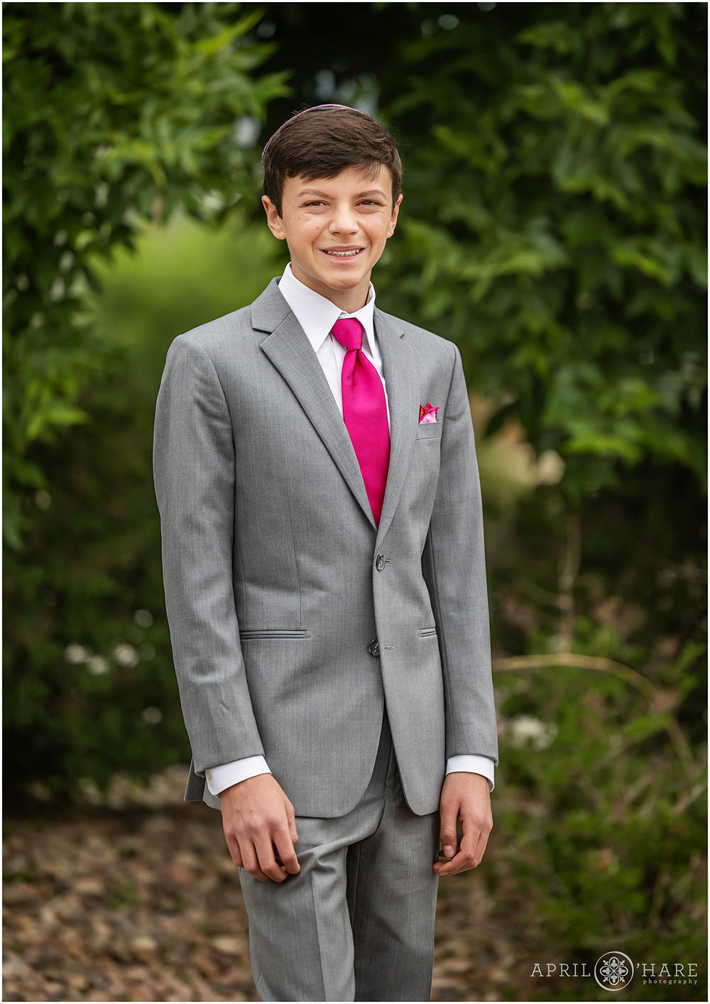 Bar mitzvah boy poses in front of some greenery in his gray suit wearing a bright pink tie at B'Nai Chaim in Lakewood