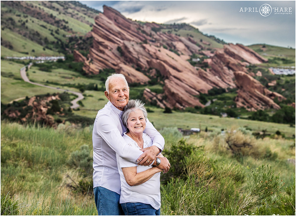 Older couple wearing white tops and blue jeans embrace at East Mount Falcon Trailhead with Red Rocks in the distance in Morrison Colorado